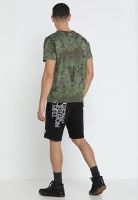 T-shirt verde camouflage con maniche corte, abbinata a pantaloni corti neri con stampa del logo bianca. L'outfit include calze scure e scarpe nere.