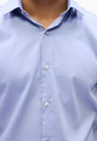 Gros plan d'une personne portant une chemise bleu clair à boutons avec col, montrant trois boutons visibles sur le devant.
