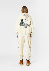 Persona vestida con una sudadera color crema y pantalones con un gráfico de dibujos animados en la parte posterior, de pie contra un fondo blanco liso.