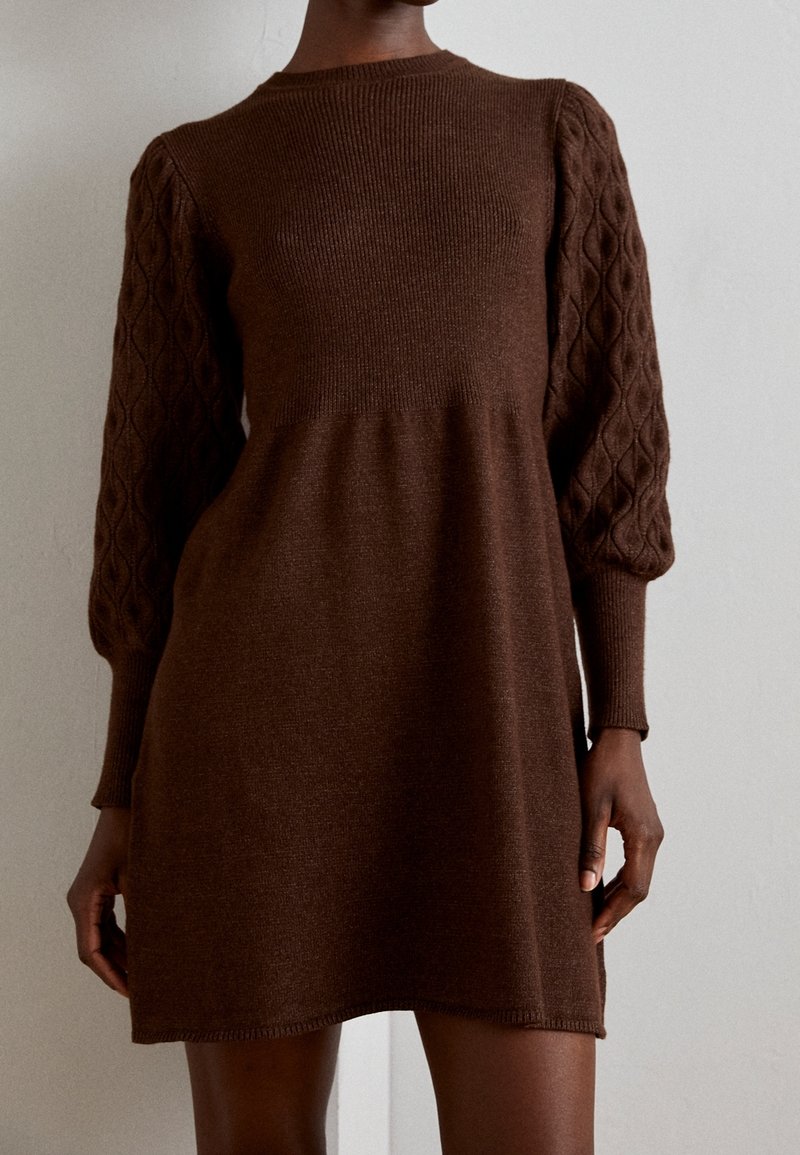 Robe en maille marron avec un corsage côtelé et des manches bouffantes en maille torsadée texturée. L'ourlet est légèrement incurvé, conçu pour une coupe décontractée.