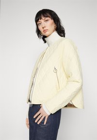 Gepolsterte gelbe Jacke mit einem Frontreißverschluss, strukturiertem Stoff und einer cropped Silhouette. Getragen über einem weißen Rollkragenpullover und dunklen Jeans.