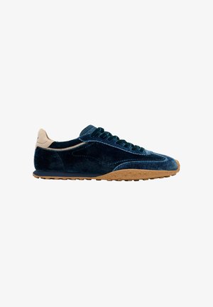Zapatilla deportiva de terciopelo azul oscuro con lengüeta en el talón beige, cordones a juego en azul oscuro y suela de goma texturizada marrón claro, vista de perfil.