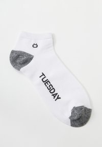 Weißer Knöchelsocke mit schwarzem Zehen- und Fersenbereich, auf der Seite mit dem Text "DIENSTAG" in Schwarz bedruckt, und einem kleinen runden Logo über dem Zehenbereich.