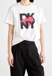 Biała bawełniana koszulka z nadrukiem graficznym z napisem "DKNY" oraz czerwoną serduszkiem. Koszulka ma krótkie rękawy i luźny krój, zestawiona z czarnymi spodniami.