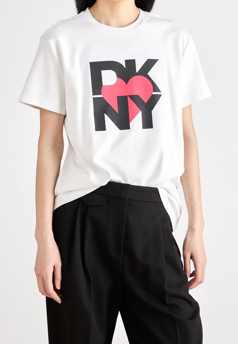 Biała bawełniana koszulka z nadrukiem graficznym z napisem "DKNY" oraz czerwoną serduszkiem. Koszulka ma krótkie rękawy i luźny krój, zestawiona z czarnymi spodniami.