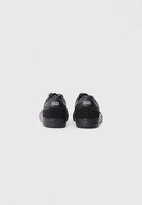 Black sneakers with a textured material, featuring a smooth sole and a logo on the heel. Rear view shows minimalistic design.