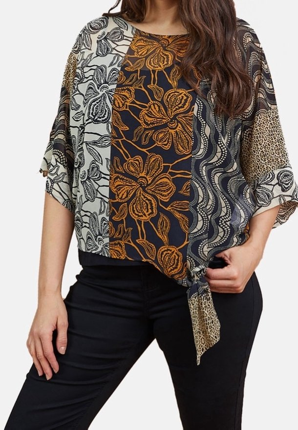 Femme portant un haut ample à manches courtes avec des motifs floraux et animaliers mélangés, noué à la taille, associé à un pantalon noir.