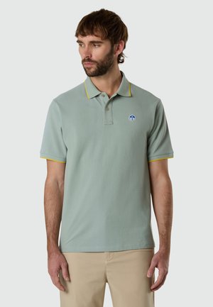Hombre con camisa tipo polo verde salvia con ribete amarillo y pantalones beige, mirando hacia la izquierda sobre un fondo liso gris claro.