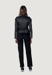 Giacca in nylon nera con silhouette aderente, dotata di dettagli sulle spalle e polsini con zip, abbinata a pantaloni in denim neri e scarpe sportive.