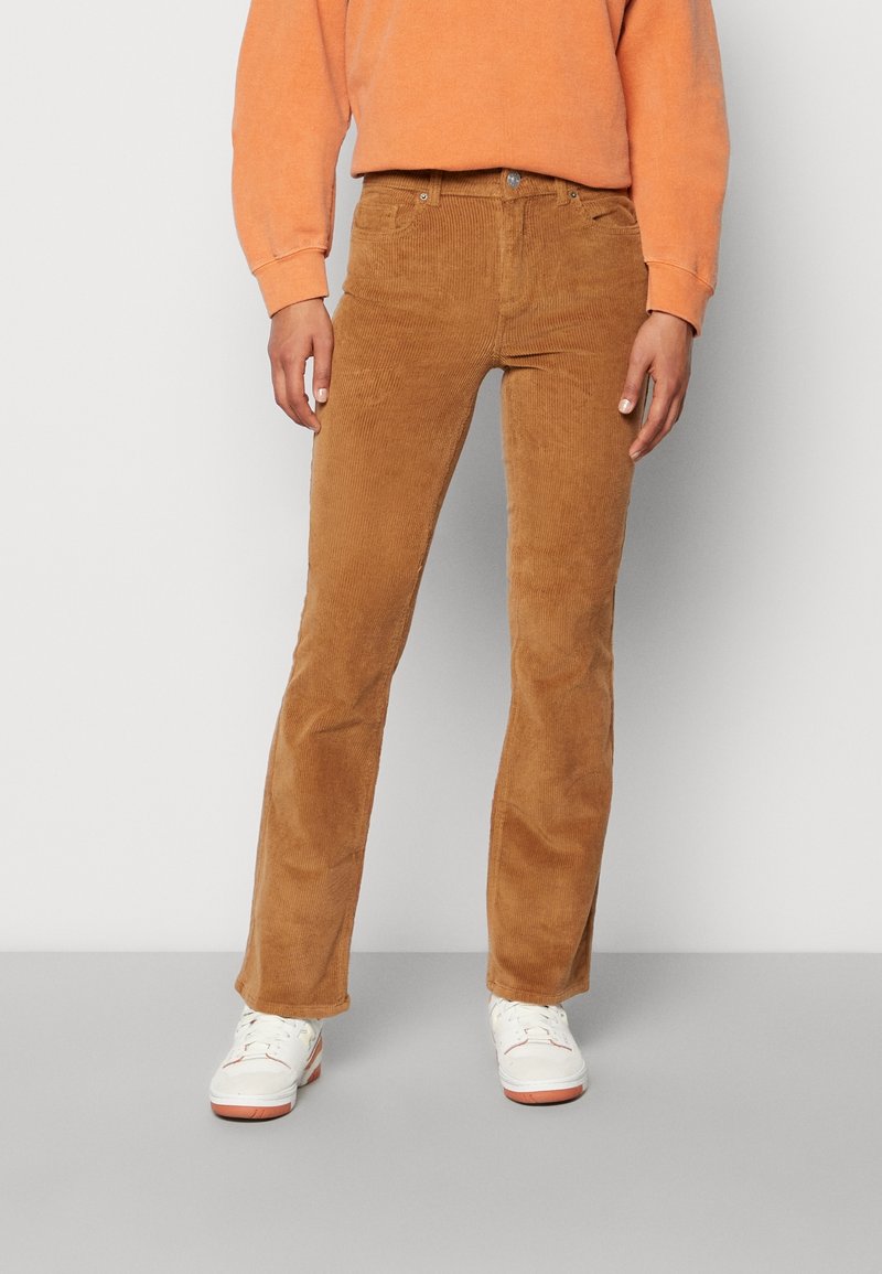 Personne portant un pantalon en velours côtelé marron clair, un haut à manches longues orange et des baskets blanches, debout devant un fond blanc uni.