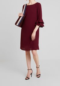 Robe bordeaux avec manches à volants, longueur au genou, texture transparente. Accessoirisée d'un sac à épaule avec une sangle rayée.
