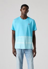 Lichtblauwe katoenen t-shirt met een tie-dye verloop, ronde hals, korte mouwen en een klein rood logo patch op de borst. Draag het met vervaagde blauwe spijkerbroek.