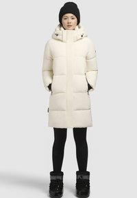 Parka blanc duveteux avec un col haut, fermeture éclair à l'avant et poches latérales, porté avec des leggings noirs et des bottes d'hiver. Bonnet en tricot noir sur la tête.