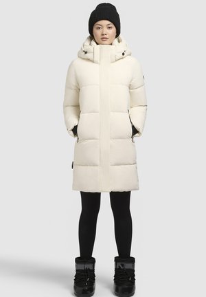 Puffige witte parka met een hoge kraag, ritssluiting aan de voorkant en zijzakken, gecombineerd met zwarte leggings en winterlaarzen. Gebreide zwarte beanie op het hoofd.