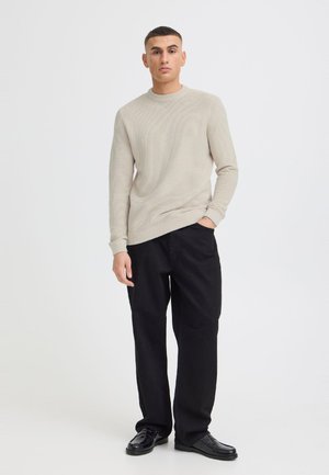 Jeune homme debout, une main dans la poche, portant un pull beige texturé, pantalon noir et chaussures en cuir noires sur un fond blanc uni.