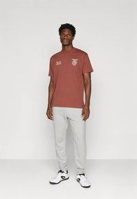 Mladý muž stojící v rustikově zbarveném tričku s potiskem, světle šedých teplácích a bílých teniskách na jednobarevném bílém pozadí.