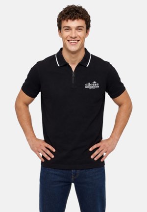 Jeune homme qui sourit, portant un polo ellesse noir avec bordure blanche et un jean bleu, les mains sur les hanches, debout devant un fond uni.