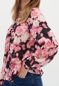 Donna che indossa una blusa floreale rosa, nera e bianca con maniche lunghe, abbinata a jeans blu, con una mano infilata nella tasca, su uno sfondo semplice.