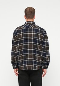 Camicia a quadri in blu scuro, marrone e bianco, con colletto a bottoni, maniche lunghe e orlo arrotondato. Il tessuto appare testurizzato e decorato.