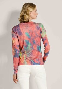 Frau mit blonden Haaren, die ein mehrfarbiges, abstraktes gemustertes Langarmoberteil und weiße Hosen trägt, mit dem Rücken zu einem schlichten Hintergrund gewandt.