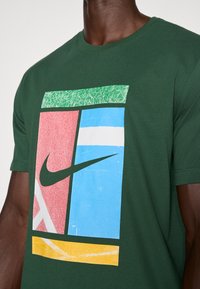 Zaļš kokvilnas T-krekls ar lielu grafisko dizainu, kas satur sarkanus, zilus, zaļus un dzeltenus laukumus, tostarp melno Nike logotipu.