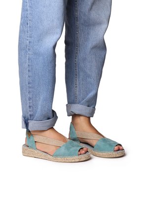 Turkoosi mokkanahasta valmistetut espadrilles-sandaalit, joissa on avoimet varpaat, elastiset hihnat ja punottu juuttipohja. Yhdistettyinä käärittyjen vaaleiden farkkujen kanssa.