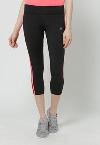 Woman wearing black Adidas cropped leggings with pink side stripes, paired with gray sneakers and a coral top, standing against a white background.