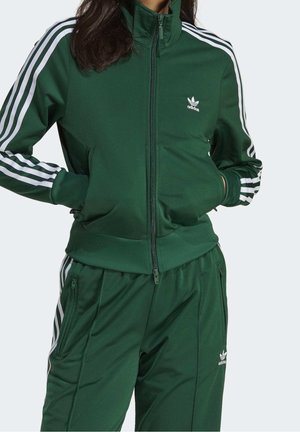 Personne portant un survêtement Adidas vert foncé avec des bandes blanches sur les manches et le pantalon, mains dans les poches de la veste, visage non visible.
