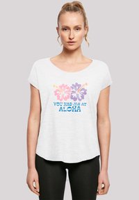 Weißes T-Shirt mit kurzen Ärmeln in lockerer Passform, mit einem grafischen Druck von rosa und lila Hibiskusblüten und blauem Text, der "DU HAST MICH BEI ALOHA ERWISCHT" sagt.