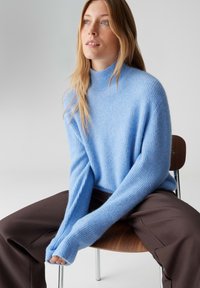 Pull bleu clair en tricot avec un col montant et une texture côtelée, assorti à un pantalon large marron foncé. Assis sur une chaise en bois.
