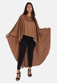 Poncho de tela marrón y fluida con mangas anchas, que presenta una textura suave y una silueta holgada, combinado con pantalones negros ajustados y sandalias de tacón.