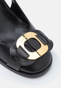 Chaussure plate en cuir noir avec bout rond, dotée d'une grande boucle décorative dorée sur la bride avant avec nom de marque embossé.