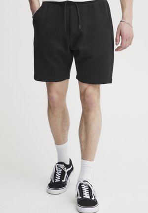 Black shorts made of textured fabric, featuring an elastic waistband with drawstring. Worn with black and white sneakers and white socks.