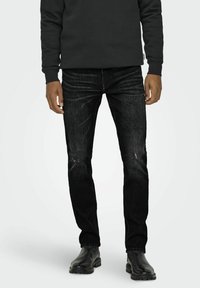 Jeans negri din denim cu o croială slim, ușor decolorate în zona genunchilor, și design cu 5 buzunare, purtate cu bocanci negri din piele și un hanorac închis la culoare.