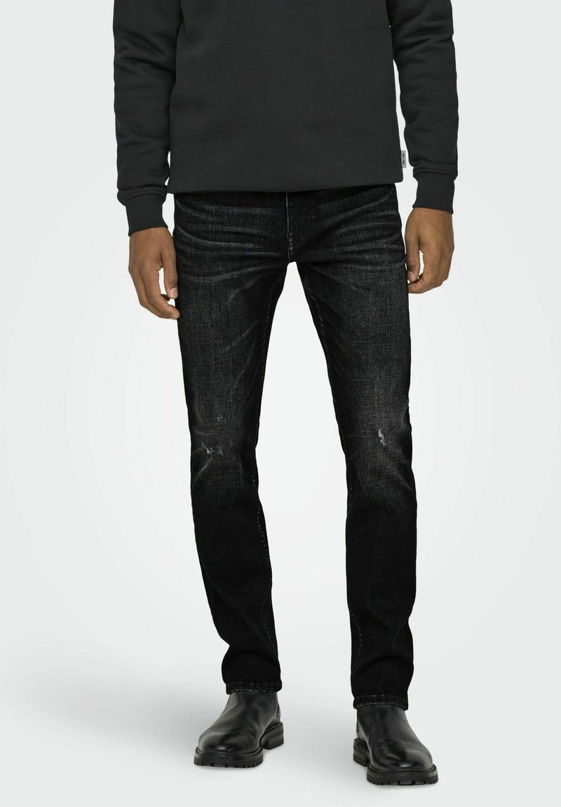 Jeans negri din denim cu o croială slim, ușor decolorate în zona genunchilor, și design cu 5 buzunare, purtate cu bocanci negri din piele și un hanorac închis la culoare.