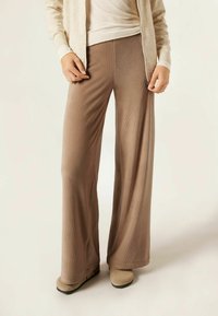 Løse ribbede bukser i lysebrun med en blød tekstur. Båret med beige fodtøj og en off-white cardigan, vist fra taljen og ned.