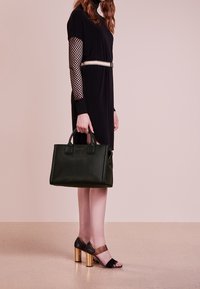 Vihreä nahkalaukku, jossa on rakenteellinen muotoilu, kaksinkertaiset kahvat ja hienovaraiset työnnökselliset yksityiskohdat, jota pidetään henkilön toimesta, joka on pukeutunut mustaan mekkoon ja korkokenkiin.