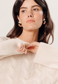 Lichtbeige kabelbrei trui met geribde manchetten en halslijn. Model draagt gouden en parel oorringen. Zachte textuur, relaxte pasvorm.