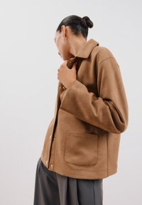 Ruskea, pehmeä kangastakki, jossa on kauluksellinen kaulus, nappisuljenta ja yksi tasku edessä. Kyynärpäähän ulottuvat hihat, rento malli.