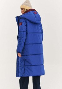 Niebieska kurtka puchowa z pikowanym wzorem, dużym kapturem oraz bordowymi akcentami na kołnierzu i rękawie. Posiada długi krój oraz zapięcia na guziki.