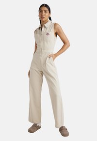 Mono de pana beige con cierre delantero, cuello corto, dos bolsillos en el pecho y bolsillos laterales. La modelo lleva unos zapatos marrones de tipo slipper y calcetines blancos.