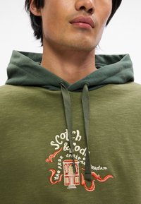 Ελιάς πράσινη κουκούλα με δίχρωμο σχέδιο, που διαθέτει ανοιχτό πράσινο επένδυση και κεντημένο λογότυπο με κόκκινες λεπτομέρειες και λεπτομέρειες χταποδιού.