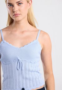 Light blue knitted camisole with scalloped neckline, adjustable straps, and a waist tie. Features ribbed texture and openwork pattern at the bottom.