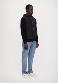 Hanorac zip-up negru și gri cu un corp texturat, mâneci uni și o buzunare stil cangur, purtat cu blugi de culoare albastră deschisă și adidași albi.