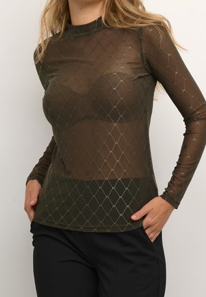 Femme portant un haut ajusté, à manches longues, transparent, vert olive avec motif losange, associé à un pantalon noir, mains dans les poches.