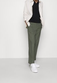 Ljust beige skjorta i jackstil över en svart polo, parat med olivgröna byxor och vita sneakers med tryckta detaljer.