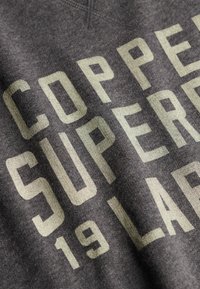 Tissu gris foncé avec de grandes lettres majuscules en crème clair formant "COPPER SUPER 19 LA", imprimées en diagonale.