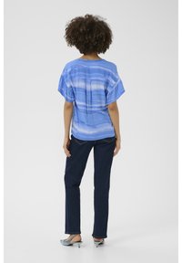 Zils rakstains īsām piedurknēm krekls ar brīvu piegriezumu, kombinēts ar tumšajiem džinsiem un gaiši ziliem augstpapēžu apaviem, rādīts no aizmugures.