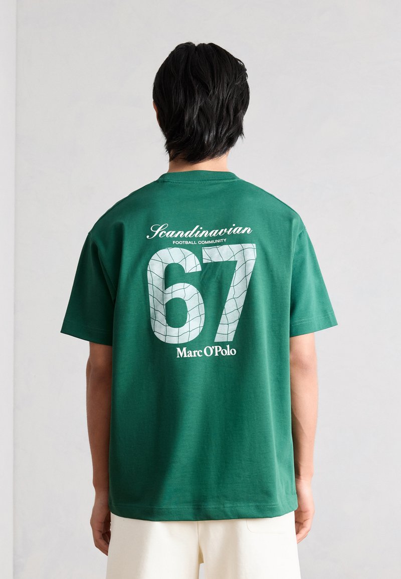 Άτομο που φοράει πράσινο T-shirt με λευκό κείμενο «Scandinavian Football Community 67 Marc O'Polo» από πίσω, μπροστά σε απλό τοίχο.