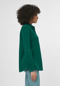 Lange mouwen groene blouse met knoopkraag, zachte textuur, ontspannen pasvorm en gebogen zoom. Gekleed met geblokte broek.